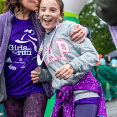 A Girls on the Run participant and her running buddy smile while proudly showing off 5K medal outdoors at the finish line