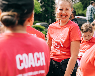 A smiling female coach in a pink t-shirt with the word "COACH" on the back looks over her shoulder. Another coach and a young girl are visible in the background. d.	Description =     A candid, instant-print style photo captures a joyful female coach in a pink t-shirt, smiling warmly. In the background, another coach and a young team mem
